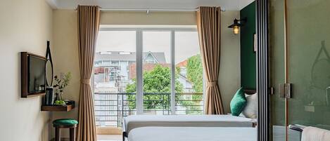 Habitación de hotel con cama, televisión, una mesita con un florero y vistas a edificios y árboles a través de amplias ventanas.