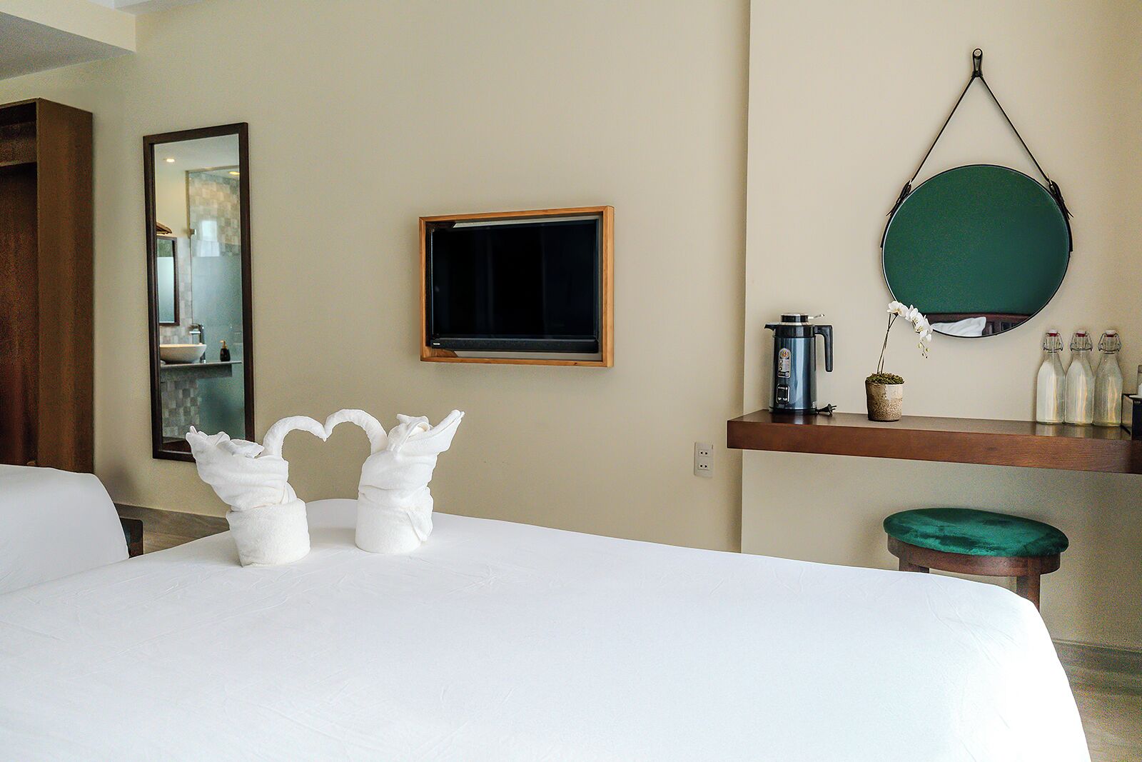A hotel room with a large bed, a flat-screen TV, a mirror, a small table with a coffee maker, and a green cushion.