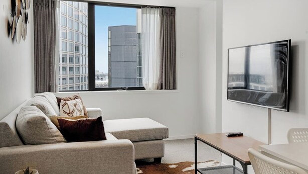 A modern living room with a sofa, a coffee table, and a TV. There is a window with curtains and a view of buildings outside. - CBD 2-Bedroom Apt near Hospital and K`Rd (Auckland CBD)