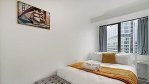 A hotel room with a bed, a window with curtains, a painting on the wall, and a view of buildings outside. - CBD 2-Bedroom Apt near Hospital and K`Rd (Auckland CBD)
