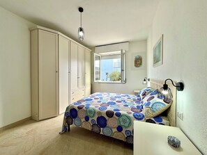 A bedroom with a bed, a wardrobe, a window, and a wall-mounted lamp. - " Family Apartment " (Tortolì)