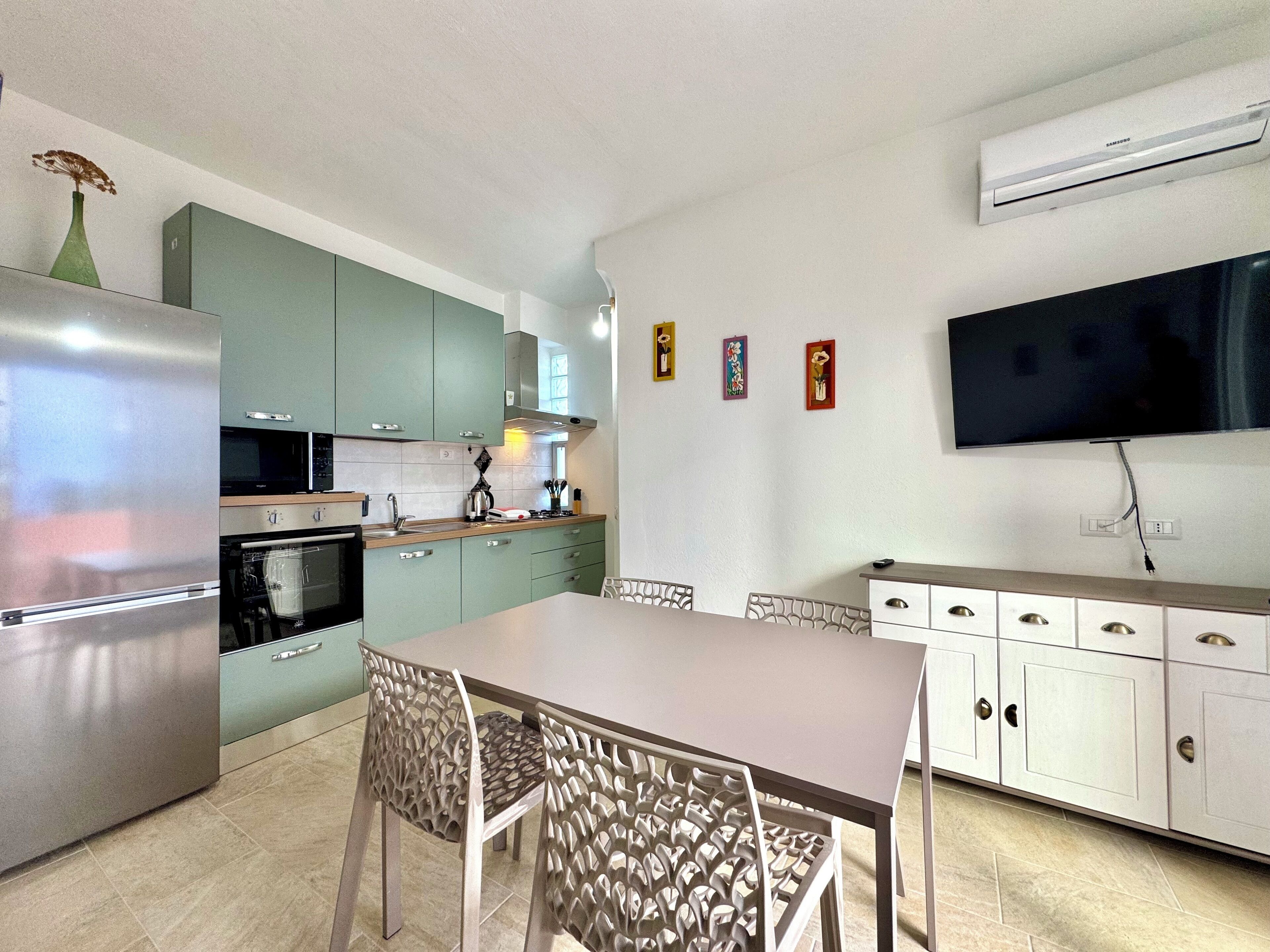 Cozinha moderna com área de jantar, destacando eletrodomésticos de aço inoxidável, armários verdes e uma TV de tela plana.