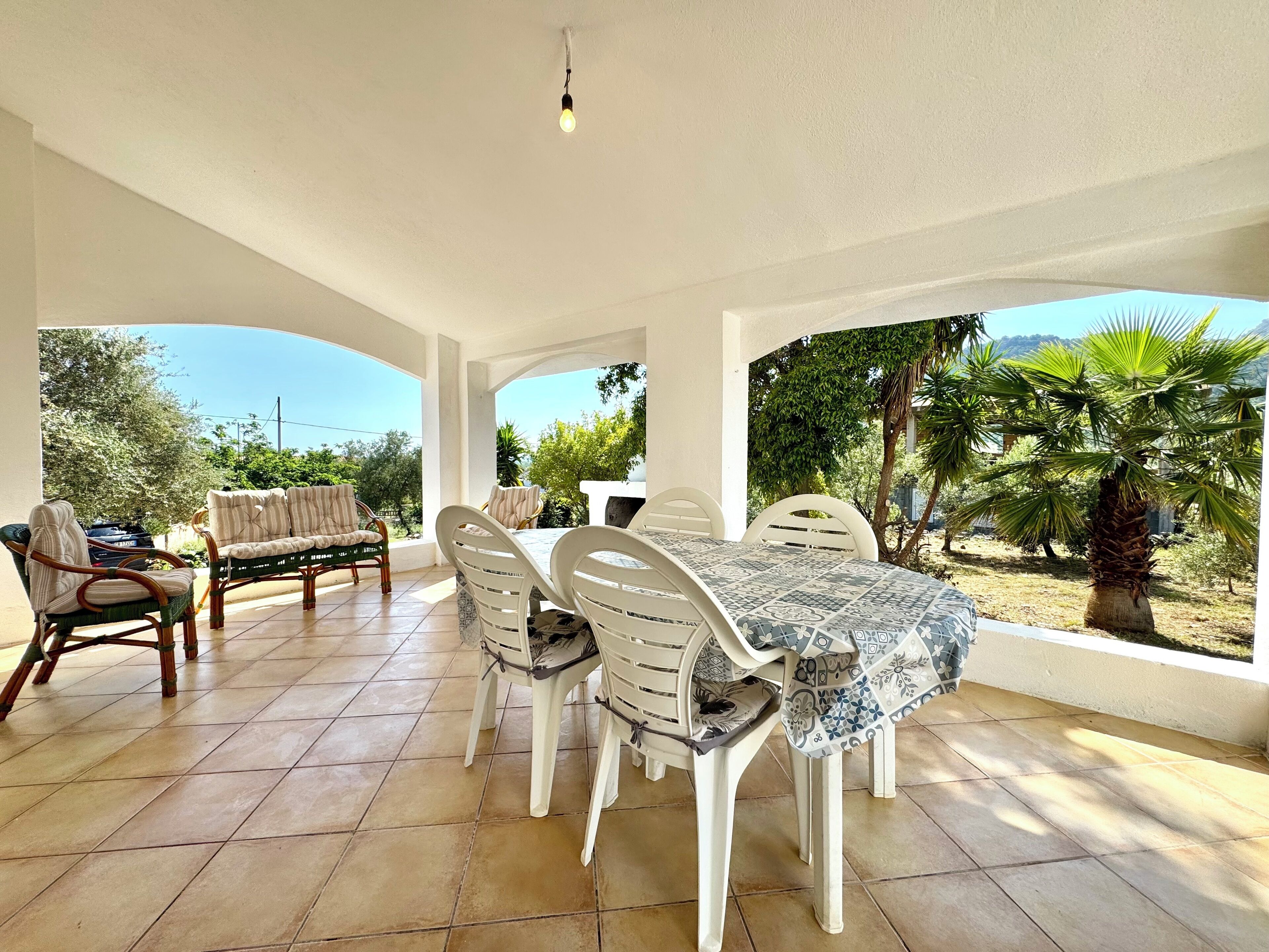 A covered patio with a dining table and chairs, a sofa, and a view of greenery and hills.