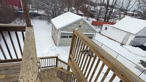 Una terraza de madera con barandilla, suelo cubierto de nieve y una casa al fondo.