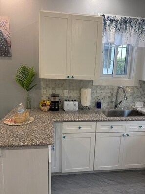 A kitchen with white cabinets, a granite countertop, a sink, a window with floral curtains, and a potted plant. - Cozy 1-bedroom house in beautiful North Palmetto Point with AC. (North Palmetto Point)