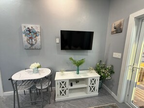 A small, well-lit room with a round table and chairs, a white cabinet with decorative plants, a mounted TV, and artwork on the walls. - Cozy 1-bedroom house in beautiful North Palmetto Point with AC. (North Palmetto Point)