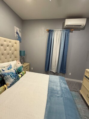 A bedroom with a tufted headboard, a bed with patterned pillows, a bedside lamp, a blue and white curtain, and a wall-mounted air conditioner. - Cozy 1-bedroom house in beautiful North Palmetto Point with AC. (North Palmetto Point)