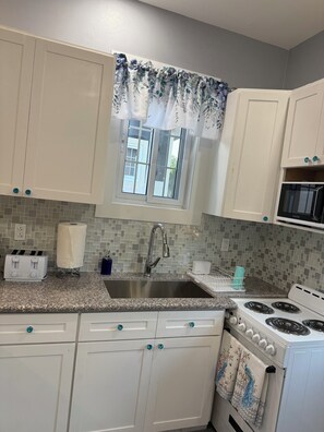 A kitchen with white cabinets, a stainless steel sink, a stove, and a window with a floral curtain. - Cozy 1-bedroom house in beautiful North Palmetto Point with AC. (North Palmetto Point)