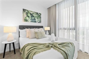 Hotelkamer met een bed, twee bedlampen, een groene deken, een schilderij en uitzicht door dunne gordijnen.