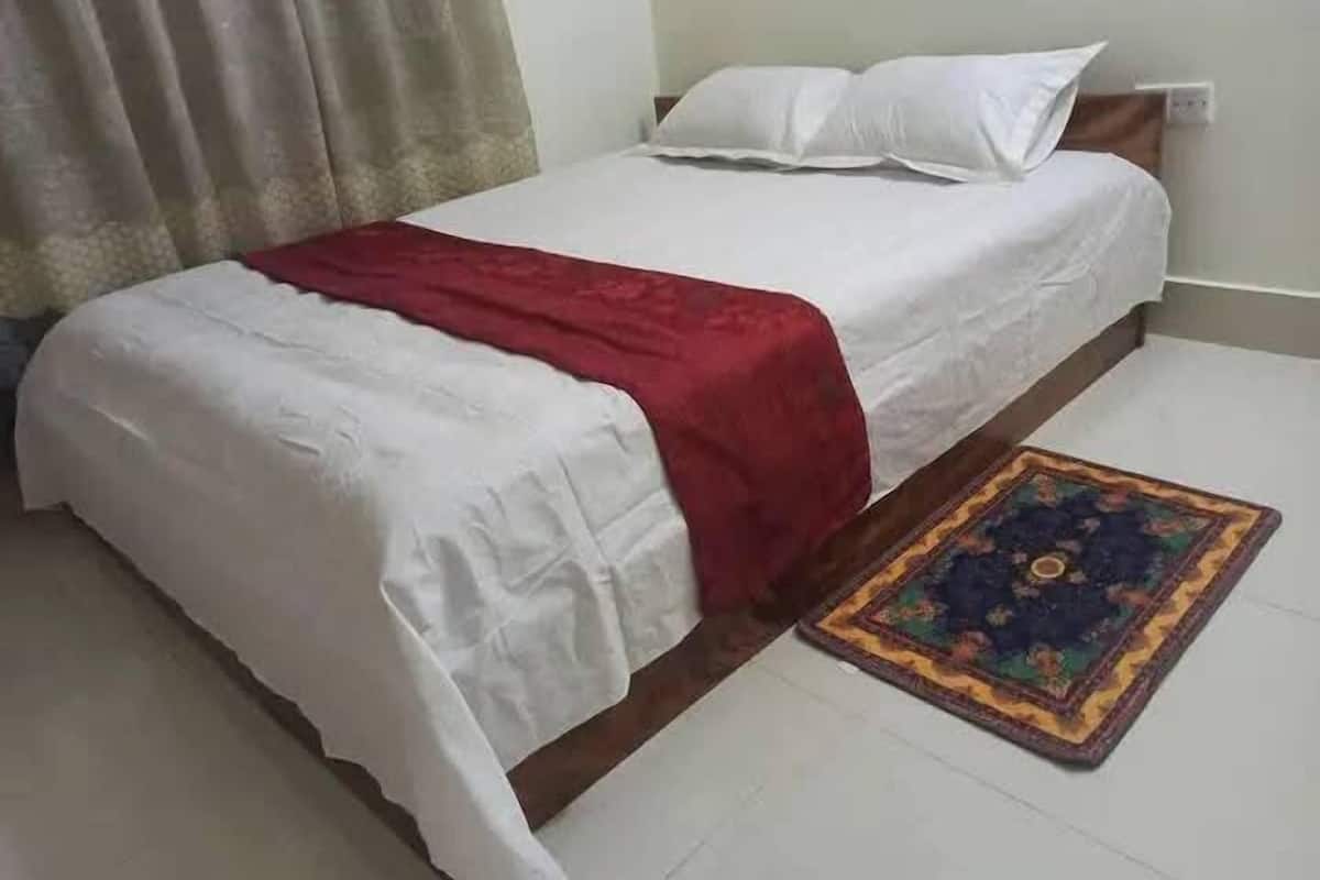 A neatly made bed with white bedding and a red blanket, a patterned rug on the floor, and a curtain partially drawn to the side.