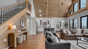 Un amplio salón con una escalera de madera, una cocina al fondo y ventanas grandes.