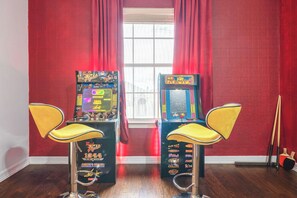 Dos máquinas de videojuegos con sillas amarillas, una ventana con cortinas rojas y una escoba apoyada contra la pared.