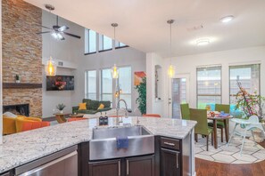 A modern kitchen with a granite countertop, stainless steel sink, and a built-in oven. There is a dining area with a table and chairs, and a living room with a sofa and a TV mounted on the wall. - Spacious, Universal Kids< 10mins, Gameroom, 18 ppl (Little Elm)