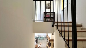 Eine Treppe mit schwarzer Geländer führt zu einer zweiten Etage. Dort ist ein Bücherregal zu sehen, während sich darunter ein Essbereich mit Stühlen und einem Tisch befindet.