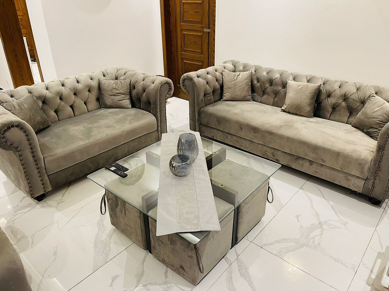 Deux canapés à accoudoirs et à dossier capitonné, ainsi qu’une table basse en verre, agrémentée d’une télécommande et d’objets décoratifs.