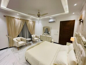 Ein Schlafzimmer mit einem Bett, zwei Sessel, einem kleinen runden Tisch, einem Schminktisch und einem Deckenventilator.