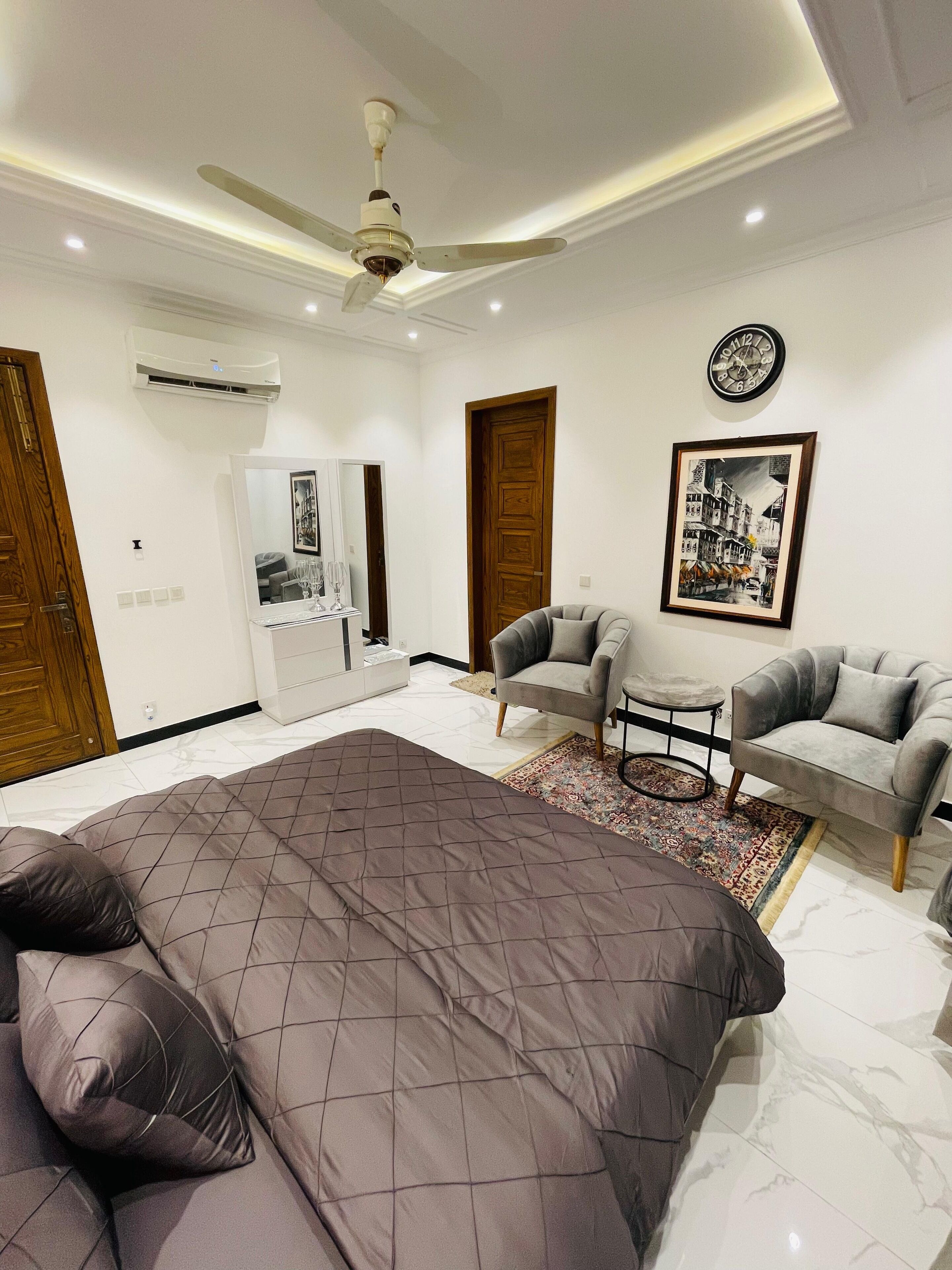 A bedroom with a large bed, a ceiling fan, a wall-mounted air conditioner, a framed picture, and a small table with a chair.