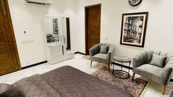A bedroom with a large bed, a ceiling fan, a wall-mounted air conditioner, a framed picture, and a small table with a chair.