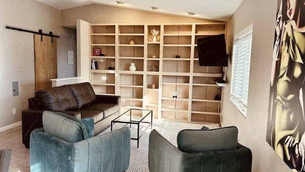 A living room with a brown leather sofa, two teal armchairs, a glass coffee table, and a built-in shelving unit with various items.