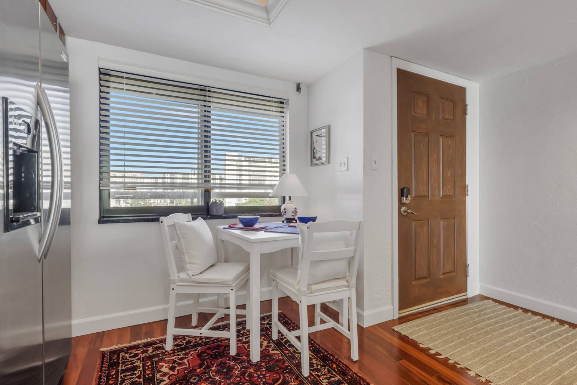 Une petite pièce avec un bureau blanc, deux chaises blanches, une fenêtre avec des stores, une porte en bois et un tapis à motifs.
