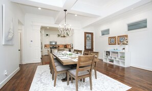 A spacious dining area with a wooden table set for six, surrounded by chairs. A chandelier hangs from the ceiling, and the room features white cabinetry and a wooden floor.