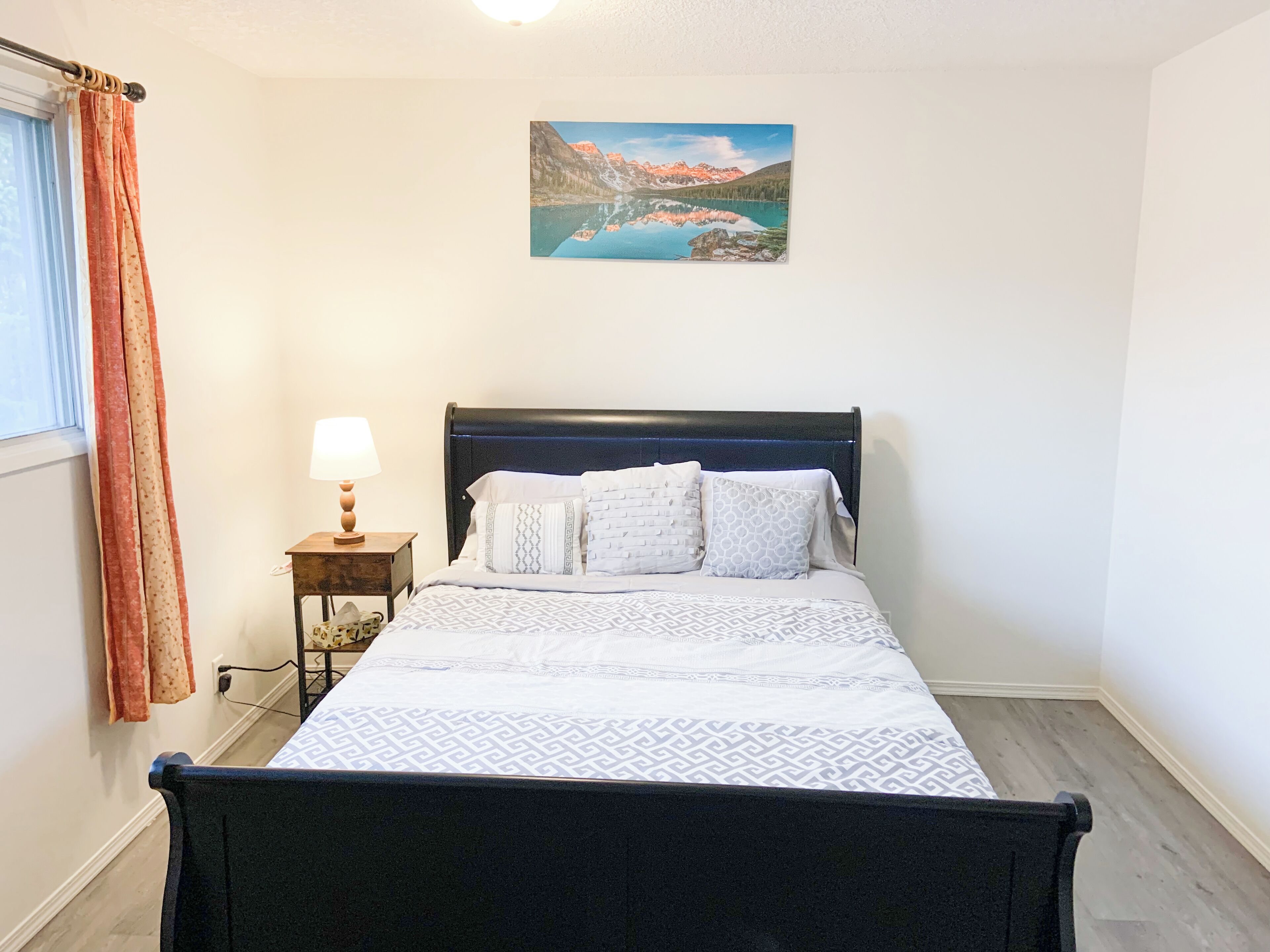 A bedroom with a bed, a nightstand, a lamp, a painting, and a window with curtains.