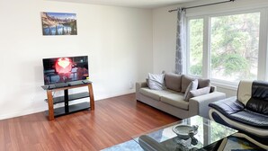 A living room with a flat-screen TV on a stand, a beige sofa, a black leather chair, a glass coffee table, and a large window with curtains. - 2 Bed rooms, 3 beds with garage for a cozy stay in Calgary (Calgary)
