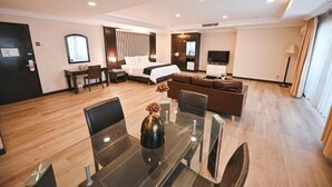 A modern hotel room with a large bed, a sofa, a dining table with chairs, a TV, and a lamp.