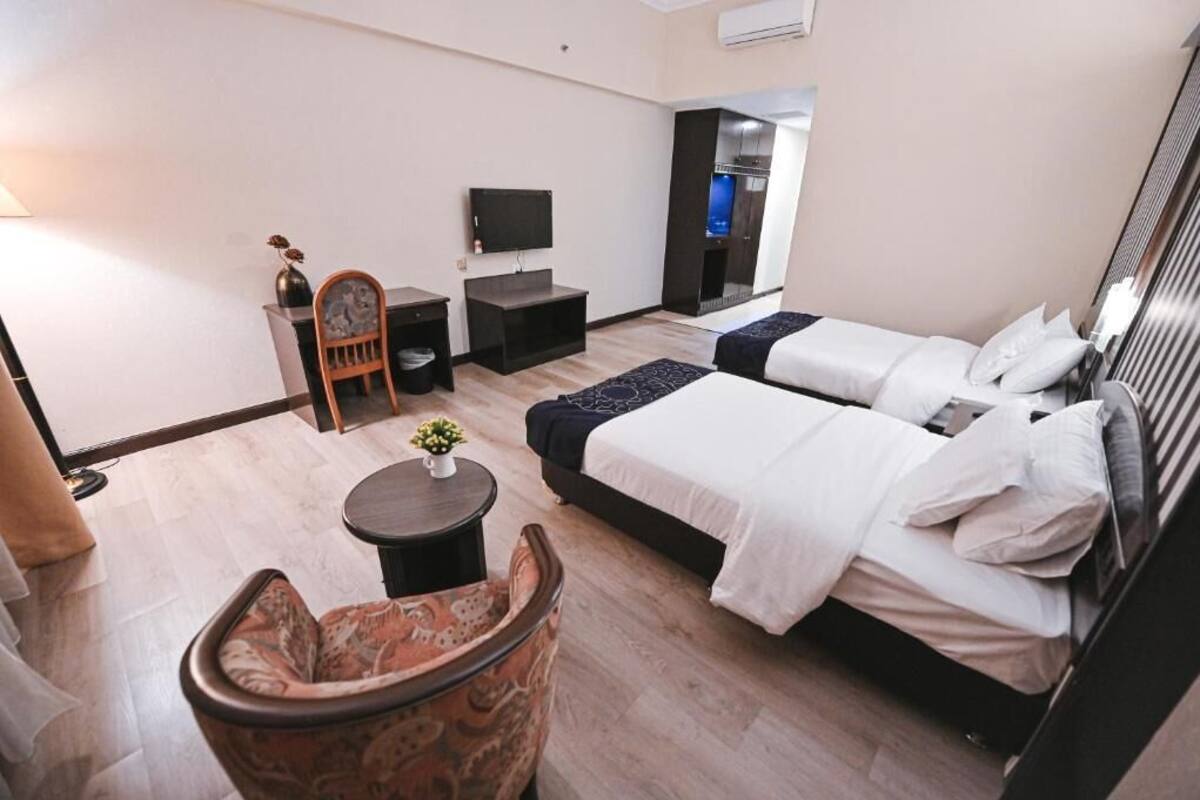 A hotel room with two beds, a TV, a small table with a vase, and a chair.