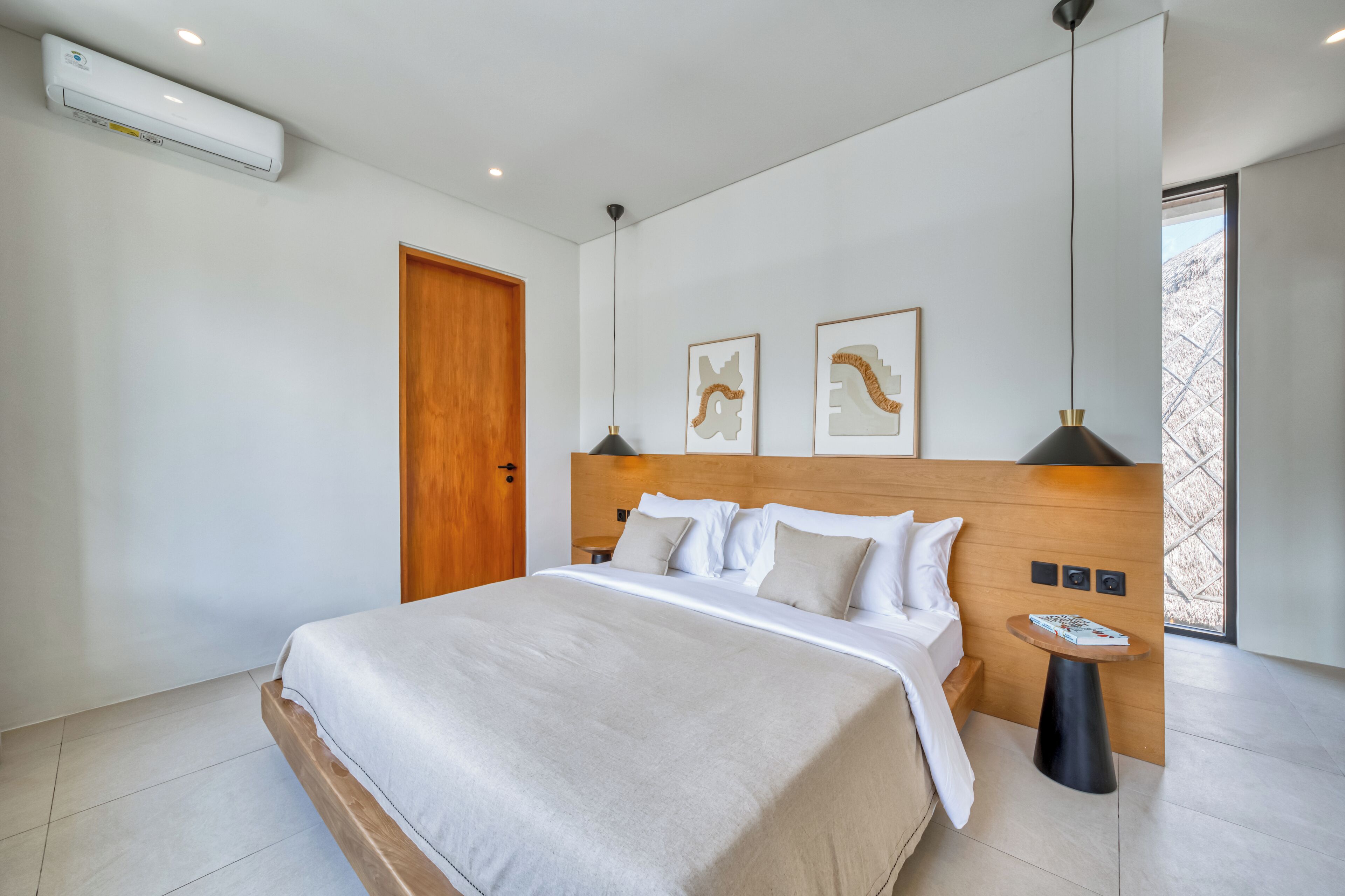 A modern bedroom with a large bed, a wooden headboard, a small side table, and a window with a view of trees.