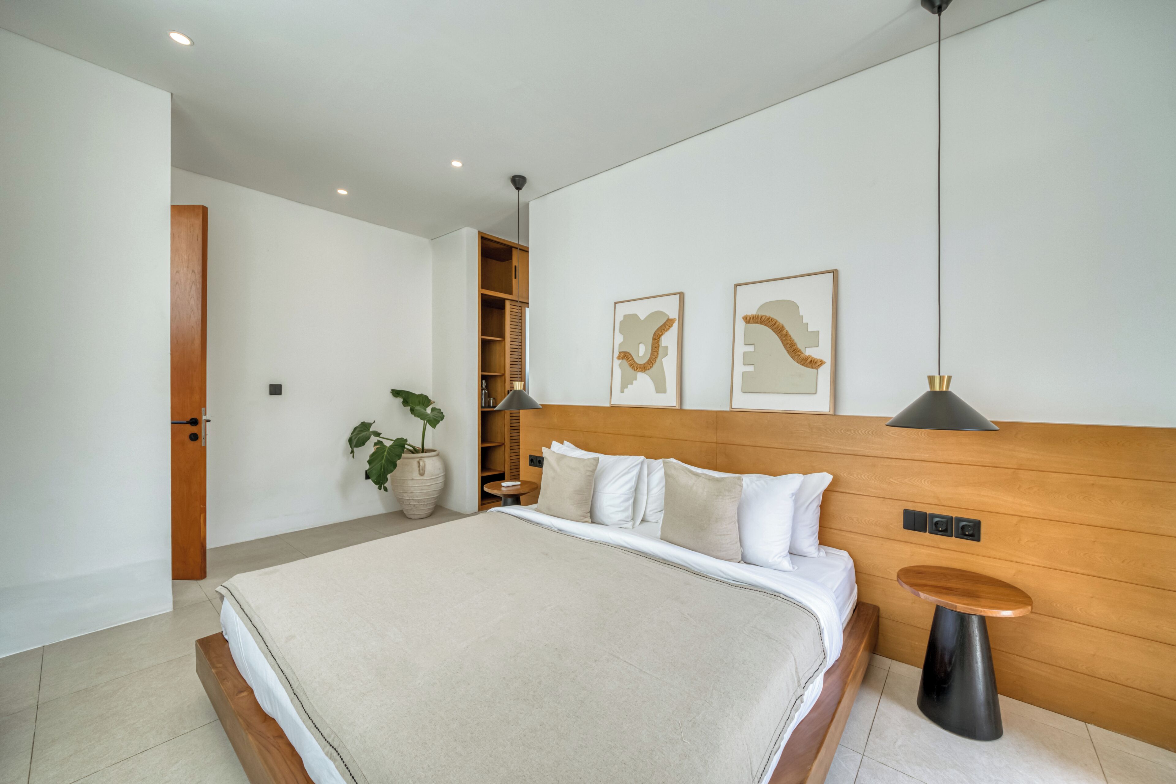 Ein modernes Schlafzimmer mit einem großen Bett, Nachttischen, einem Holz-Kopfstück, einer Topfpflanze und Bildern an der Wand.