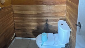 Een kleine badkamer met houten lambrisering, een wit toilet, een blauwe emmer en een zichtbare elektriciteitsuitlaat aan het plafond.