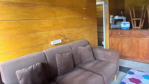 A room with a brown leather sofa, a quilted rug, wooden walls, and a bar area with dishes. - Quen'z Cabin (Kejajar)
