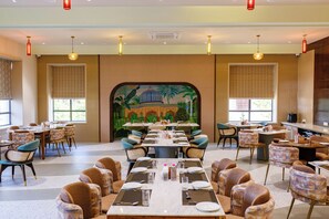 Een ruime eethoek met meerdere tafels en stoelen, een muurschildering met tropische en architectonische details, en een modern plafond met inbouwverlichting.