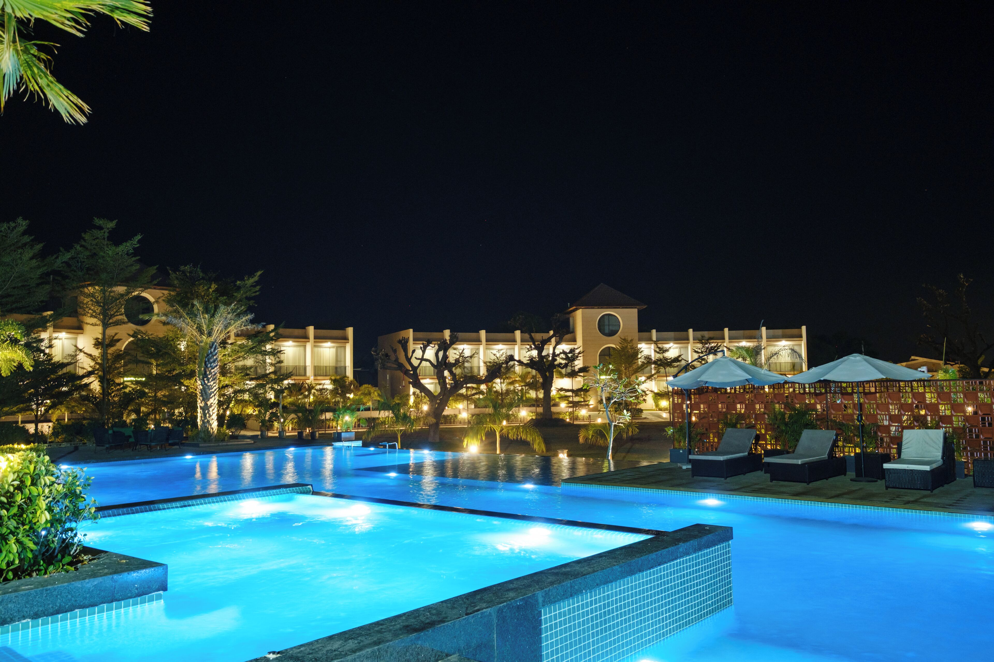 A nighttime swimming pool with lounge chairs and umbrellas, surrounded by landscaped greenery and a building with a clock tower.