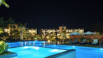 A nighttime swimming pool with lounge chairs and umbrellas, surrounded by landscaped greenery and a building with a clock tower.