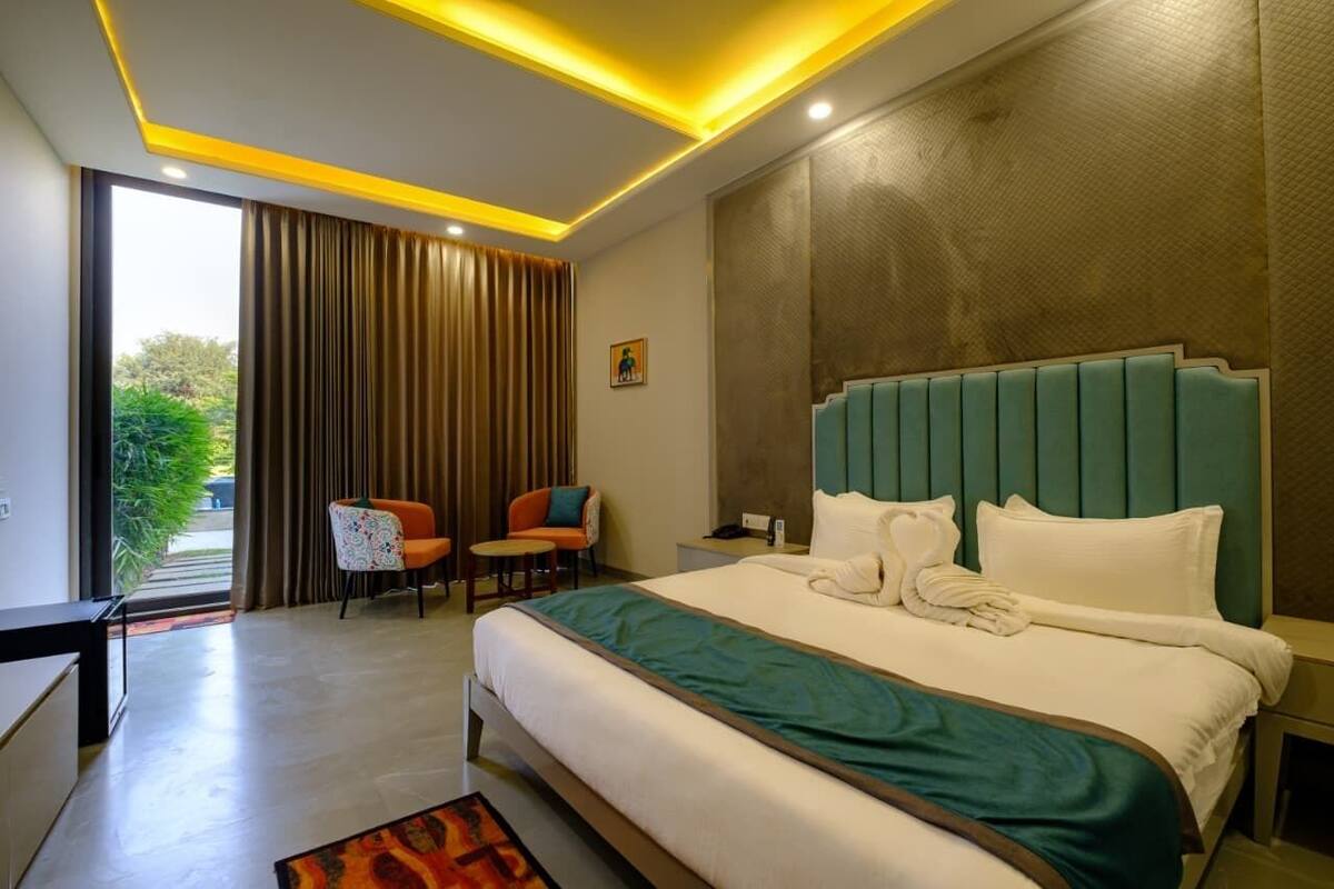 A modern hotel room with a large bed, a desk, and a chair. There is a view of greenery outside the window.