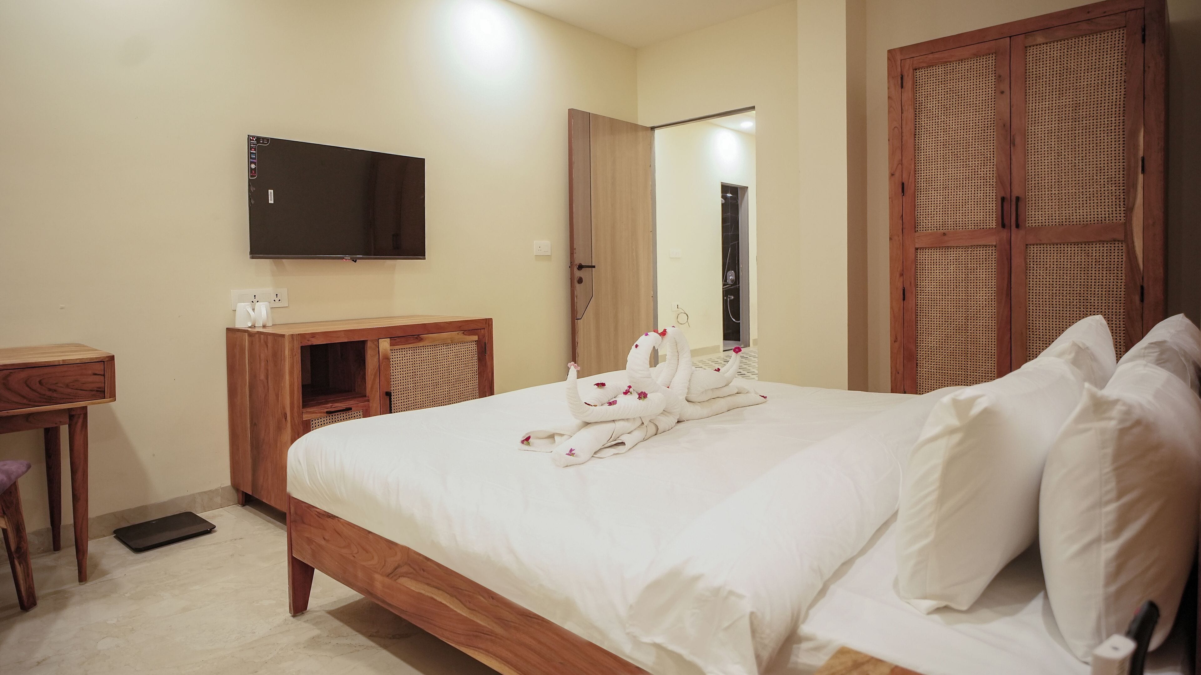 Una cama bien hecha con sábanas blancas y una toalla decorativa, una mesita de noche de madera con televisor de pantalla plana, una silla de mimbre y un armario de madera con puerta trenzada.