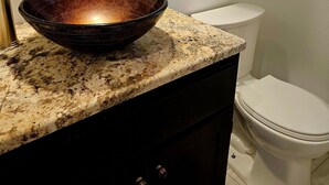 A bathroom with a granite countertop, a dark bowl sink, a white toilet, and a white shelf with a cross and a container. - Family- and Pet-Friendly Downtown Home, Close to Everything (Stevensville)