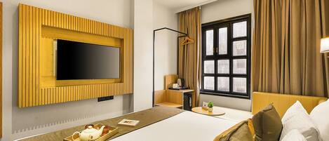A hotel room with a large flat-screen TV mounted on the wall, a bed with pillows, a bedside table with a lamp, and a window with curtains.