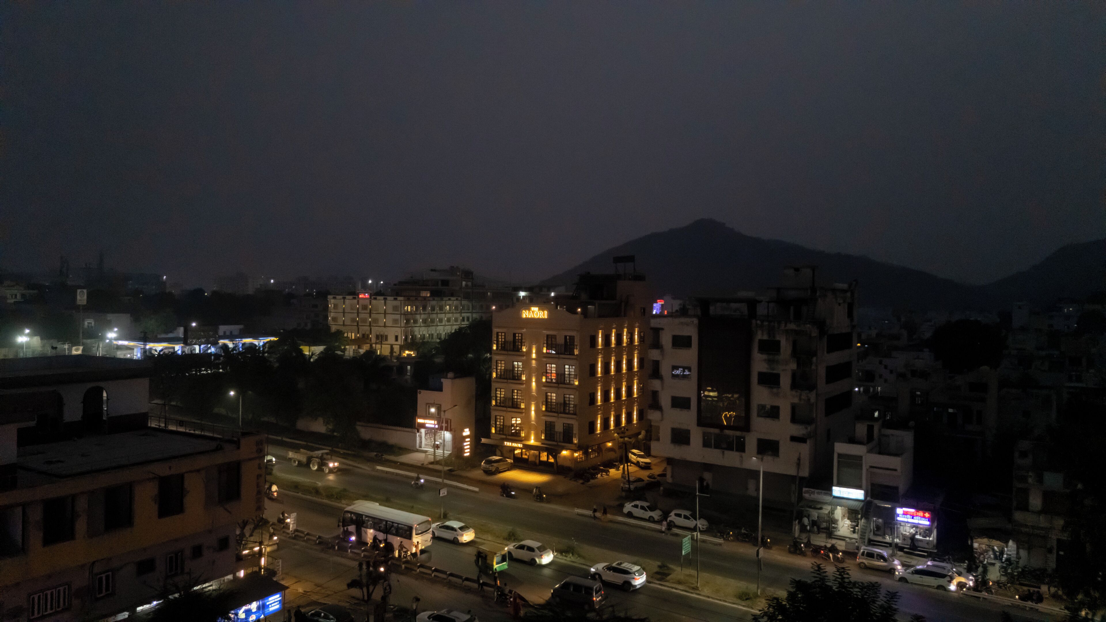 Paesaggio urbano notturno con un edificio di rilievo con la scritta 'NAORI' al centro, circondato da altri edifici e una montagna sullo sfondo.