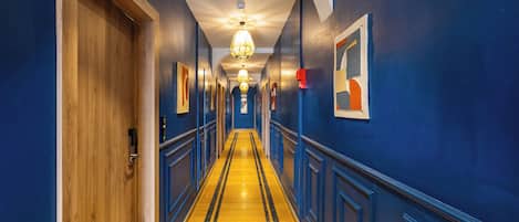 Un pasillo de hotel con una puerta de madera a la izquierda, piso a rayas amarillo y negro, y paredes azules con cuadros enmarcados.