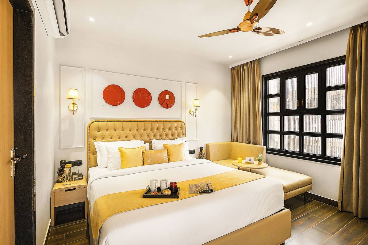 A hotel room with a large bed, a sofa, a ceiling fan, a window with curtains, and wall-mounted artwork.