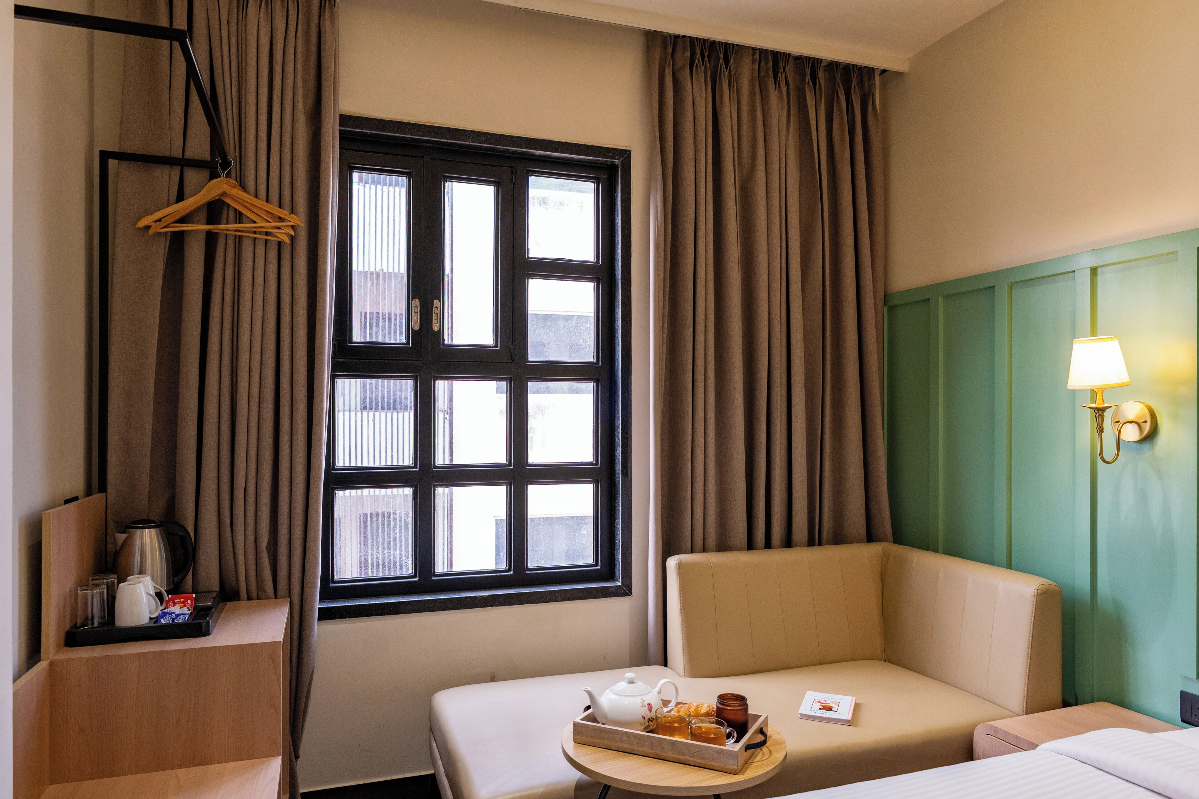 Habitación de hotel con cama, sofá, ventana con cortinas, mesa con bandeja de desayuno y una lámpara de pared.