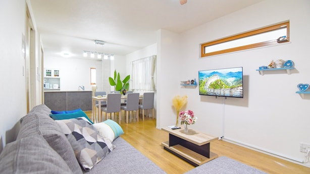 Una sala de estar moderna con un sofá gris, un comedor con sillas y una mesa, un televisor montado en la pared y un ventilador de techo.