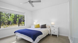 A bedroom with a bed, two bedside tables, a ceiling fan, and a window with a view of trees. - Allawah in North Bendalong (Bendalong)
