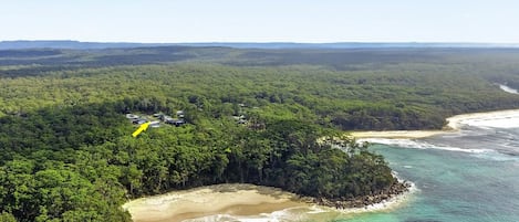 An aerial view of a coastal area with dense forest, a sandy beach, and a small settlement with buildings.
