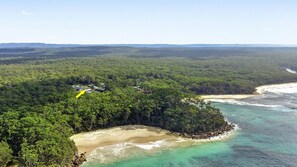 Luftaufnahme einer Küstenlandschaft mit dichtem Wald, einem Sandstrand und einer kleinen Siedlung mit Gebäuden.