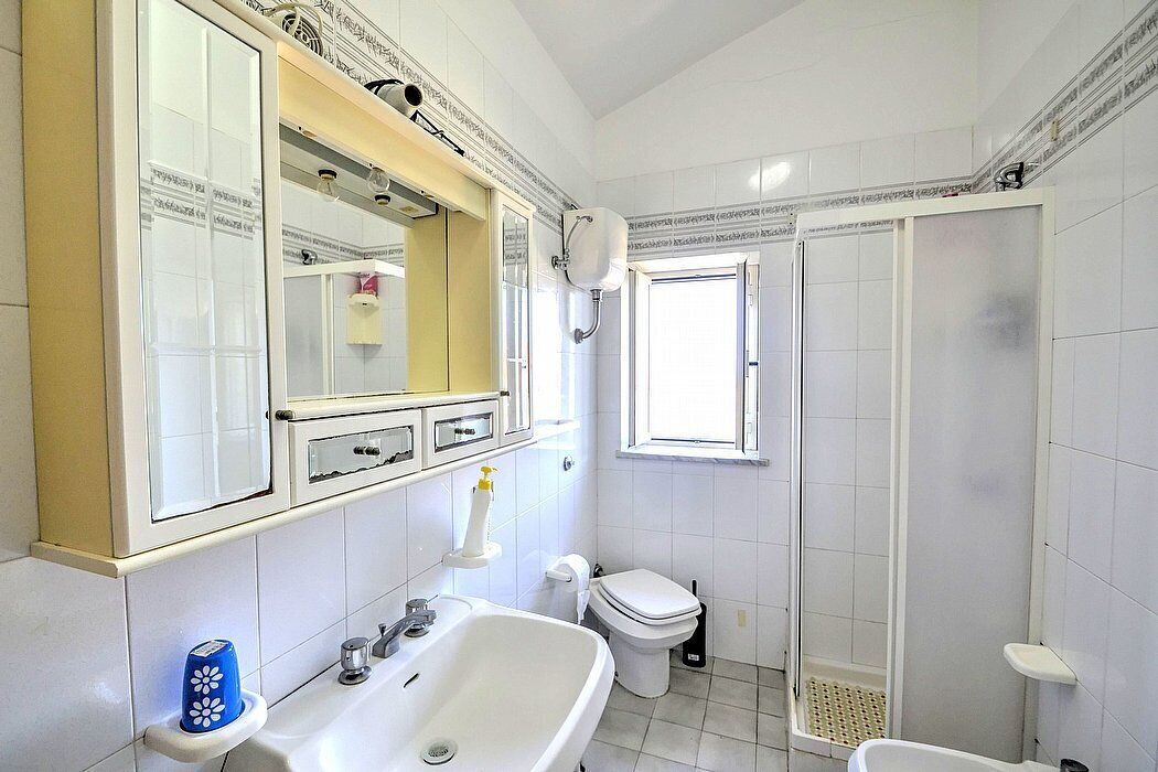 Une salle de bain avec un lavabo, des toilettes et une douche, le tout de couleur blanche. Un miroir se trouve au-dessus du lavabo et une fenêtre au-dessus des toilettes.