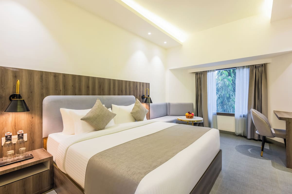 A modern hotel room with a large bed, bedside lamps, a small table with a fruit bowl, a desk, and a window with curtains.